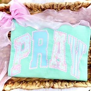 May include: A pale green t-shirt featuring the word "PRAY" in appliqued blue and floral fabric. The letters are edged with pink scalloped trim. A pink ribbon and sheer fabric are draped over a woven basket.