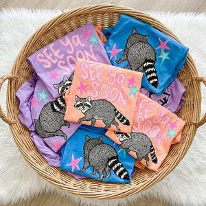 May include: A wicker basket overflowing with t-shirts. Each shirt displays a raccoon illustration and the words "See Ya Soon". The shirts are in blue, orange, and purple, with star designs. The basket sits on a white fluffy surface.