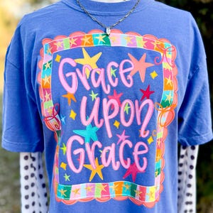 May include: A blue t-shirt featuring the phrase "Grace Upon Grace" in pink script, framed by colourful stars and a decorative border. The shirt is layered over a white and black polka dot long sleeve shirt.
