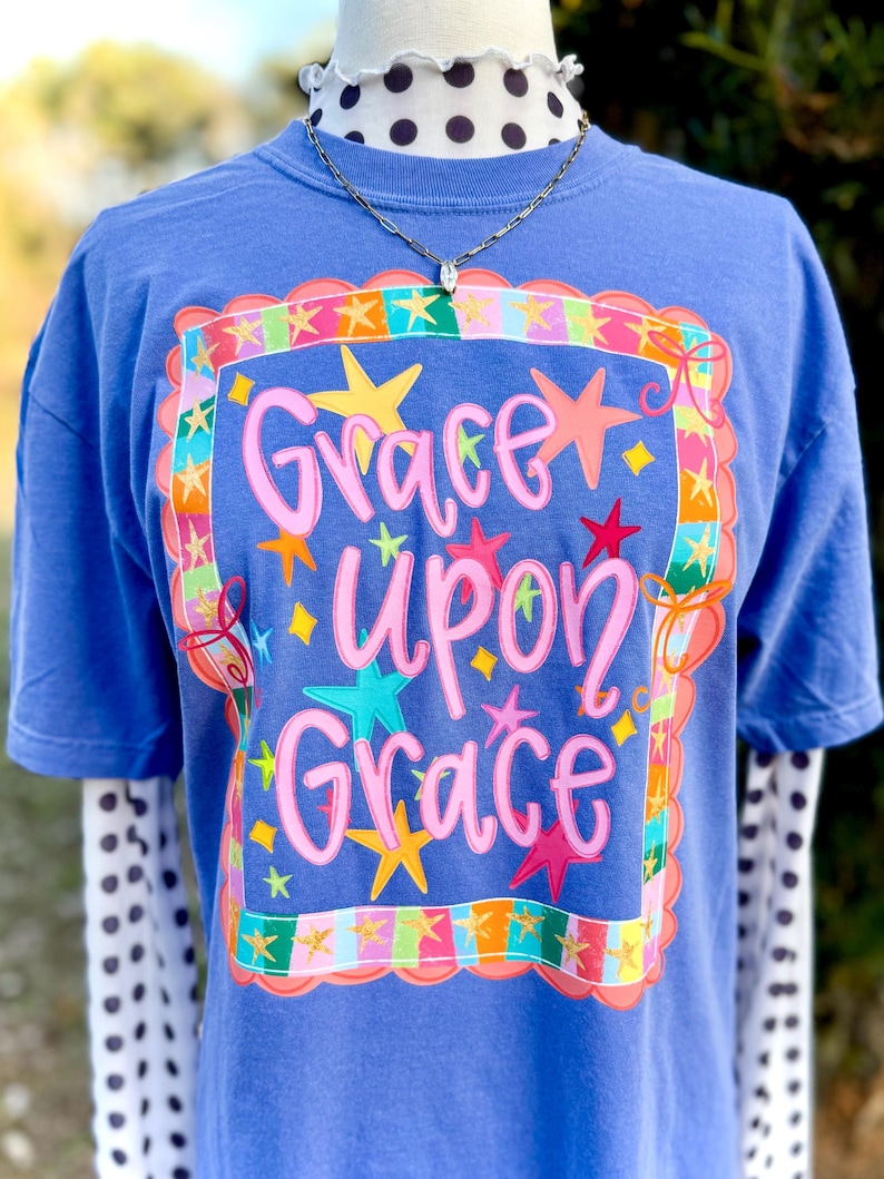 May include: A blue t-shirt featuring the phrase "Grace Upon Grace" in pink script, framed by colourful stars and a decorative border. The shirt is layered over a white and black polka dot long sleeve shirt.