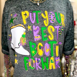 May include: A gray t-shirt with the phrase "PUT YOUR BEST BOOT FORWARD" in yellow, green, and purple. A white boot with a colorful tassel is on the left. The shirt is on a mannequin with layered necklaces.