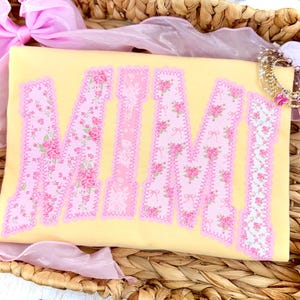 May include: A yellow rectangular item with the word "MIMI" in pink floral letters. The letters have a scalloped edge and are filled with a pink rose pattern. The item is surrounded by pink ribbons and a decorative crown.