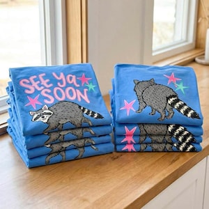 May include: Piles of blue t-shirts featuring a raccoon design and the words "SEE Ya SOON". The raccoon is grey with a striped tail, and the text is pink. The shirts are stacked on a wooden surface.