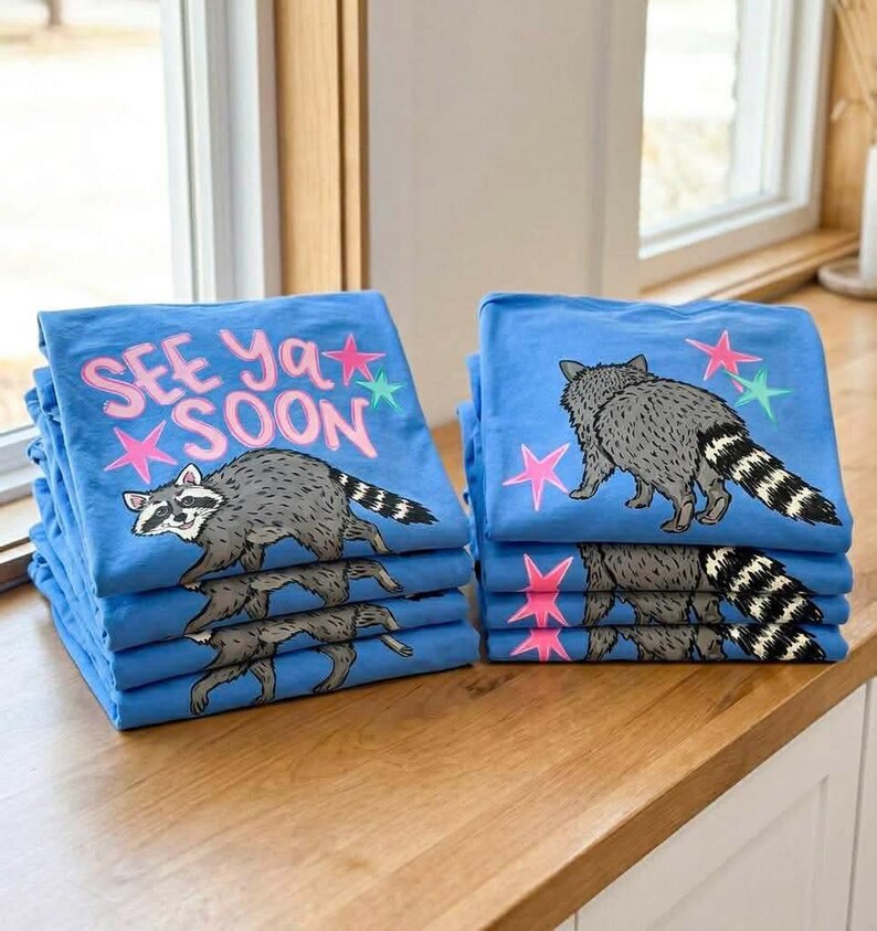 May include: Piles of blue t-shirts featuring a raccoon design and the words "SEE Ya SOON". The raccoon is grey with a striped tail, and the text is pink. The shirts are stacked on a wooden surface.