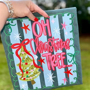 May include: A Christmas-themed square pillow with the phrase "OH Christmas TREE..." in pink script. The design includes a Christmas tree, stars, and snowflakes on a light blue striped background, framed by a green border.