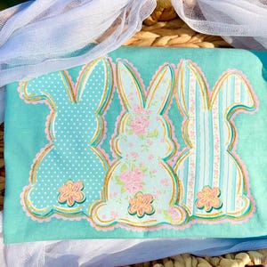 May include: A turquoise fabric with three bunny silhouettes. The bunnies have different patterns: polka dots, floral, and stripes. Each bunny is outlined in gold and pink, with a flower at the bottom. The fabric is decorated with white tulle.