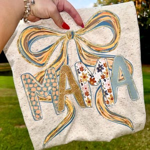 May include: A light grey t-shirt featuring the word "MAMA" in decorative lettering. The "M" is patterned with pumpkins, the first "A" with floral designs, and the second "A" in a light blue. A large bow with gold glitter accents and blue and orange details is above the word.