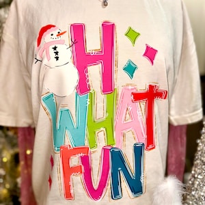 May include: Off-white t-shirt with a colorful graphic that says "Hi What Fun." The design includes a snowman wearing a red hat and pink scarf, and colorful diamond shapes. The letters are outlined in gold.