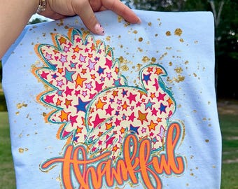 May include: A light blue t-shirt with a colourful turkey design filled with stars. The word "thankful" is written in orange script with a gold outline. Gold glitter accents the shirt. A festive design.