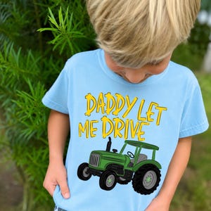 Pode incluir: T-shirt azul claro com a frase "DADDY LET ME DRIVE" em letras amarelas. Uma ilustração de um trator verde está impressa abaixo do texto. A camisa é feita de um material macio.