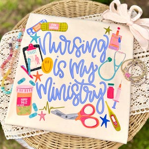 May include: Cream-colored t-shirt with the phrase "Nursing is my Ministry" in blue script. The design includes medical-themed illustrations like a stethoscope, scissors, and pills. Additional text includes "For Oopsies" and "Happy Pills".