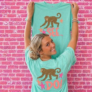 Puede incluir: Una camiseta turquesa clara con un diseño de mono marrón, estrellas rosas y el texto "SEE..." en la parte delantera. La parte trasera de la camiseta tiene el mismo diseño de mono con el texto "DO!". La camiseta se muestra contra una pared de ladrillos rosas.