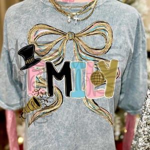 May include: A light blue t-shirt with the word "FAMILY" in a colorful design. The word is set against a large bow with gold, pink, and blue accents. A top hat, party horn, and disco ball are also featured.