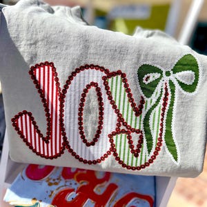 May include: A light gray sweatshirt with the word "JOY" embroidered in red and green. The letters are filled with red and white stripes, green and white stripes, and a green bow.