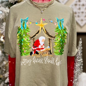 May include: Olive green t-shirt with a Christmas Nativity scene featuring Santa Claus kneeling before baby Jesus. The design includes a gold star, Christmas trees, and the text "every knee shall bow."