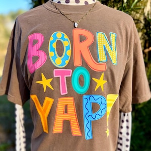 以下が含まれることがあります： カラフルなバブルスタイルの文字で「BORN TO YAP！」と書かれた茶色のTシャツ。シャツには金のネックレスが飾られており、黒い水玉模様の白い長袖シャツの上に重ね着されています。