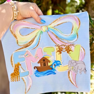 May include: A light blue shirt with a large, pastel-colored bow design and the name "EMILY" in colorful letters. The design includes a giraffe, ark, monkeys, and elephants.