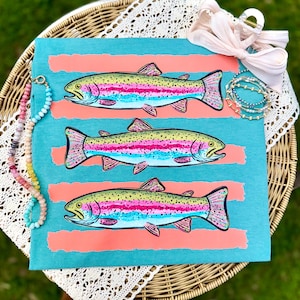Può includere: Una borsa tote turchese con tre illustrazioni di trote iridee. I pesci sono raffigurati con dettagli rosa, gialli e blu, su strisce orizzontali color corallo. La borsa è accessoriata con una collana di perline e un elastico per capelli rosa.