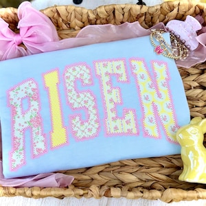 May include: A light blue t-shirt with the word "RISEN" in large, floral-patterned letters. The letters are outlined in pink and yellow. The shirt is in a woven basket with a pink bow and a yellow bunny figurine.