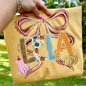 May include: Mustard yellow t-shirt with the name "LELA" in appliqued letters. The letters have various patterns, including pumpkins and plaid. A large bow design with gold glitter accents is at the top. A pink pumpkin graphic is on the left side.