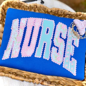 Könnte beinhalten: Ein königsblaues T-Shirt mit dem Wort "NURSE" auf der Vorderseite. Die Buchstaben sind in einer Mischung aus Blumen- und Tupfenmustern gehalten, umrandet in Rosa. Eine kleine Blumen-Haarspange und Armbänder befinden sich auf dem Shirt.
