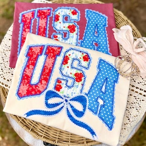 May include: Two t-shirts with the word "USA" appliqued on them. One is cream-colored with a blue bow, the other is maroon. The letters are red floral and blue polka dot patterns with a scalloped edge.