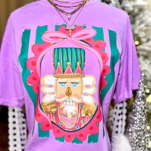 May include: A lavender t-shirt featuring a colorful illustration of a nutcracker. The design includes a pink bow, a gold crown, and a pink and green frame. Vertical green stripes are on the shirt. The shirt is displayed on a mannequin.