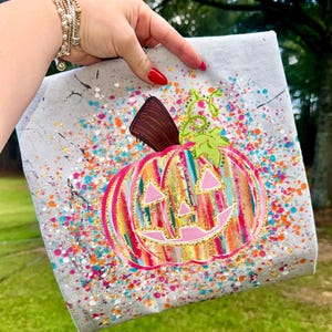 May include: A gray canvas with a colorful, painted pumpkin design. The pumpkin is pink, yellow, and orange with a brown stem and green vines. The background has colorful paint splatters and gold accents.