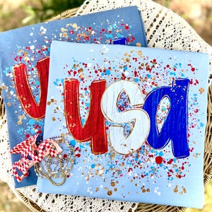 May include: Two light blue t-shirts with the word "USA" in red, white, and blue, with gold accents and paint splatter. A red, white, and blue bow and a gold necklace are in the foreground. The shirts are on a woven basket.
