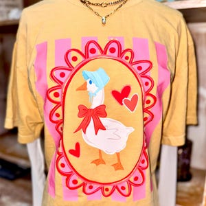 Peut inclure: T-shirt jaune moutarde avec une oie de dessin animé portant un bonnet bleu et un nœud rouge. L'oie est à l'intérieur d'un cadre ovale rose et rouge avec deux cœurs rouges. Des rayures verticales roses flanquent le cadre.