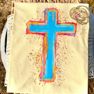 May include: A light beige canvas features a large, hand-painted cross in vibrant colors. The cross is primarily turquoise, outlined in pink, orange, and red. Gold flecks are scattered around the cross. Several gold bracelets with a pink charm are in the upper right corner.