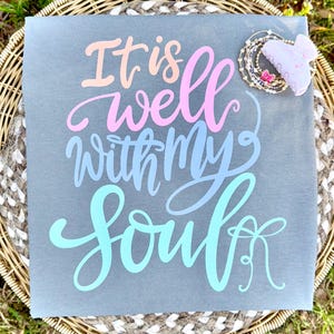 Peut inclure: Un panneau carré gris avec l'inscription "It is well with my soul" dans des couleurs pastel. Le panneau est orné d'une collection de bracelets et d'une pince à cheveux.