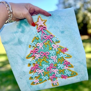 May include: A Christmas tree design embroidered on a light blue fabric. The tree is decorated with pink and blue bows, a gold outline, and a pink star on top. The fabric has a watercolor effect.