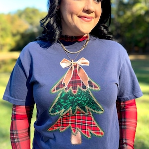 May include: Navy blue t-shirt with a Christmas tree applique. The tree is made of red and green plaid fabric, with a brown bow and gold trim. The person is wearing a red plaid long sleeve shirt underneath.