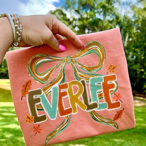 May include: A peach-colored t-shirt featuring the name "EVERLEE" in a colorful, layered font. The design includes a large bow with teal, gold, and brown accents, along with autumn leaf details. The shirt is held up against a backdrop of green grass and trees.