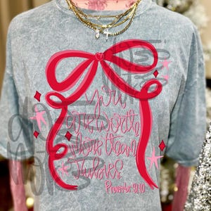 Può includere: T-shirt grigio con un grande fiocco rosso e la scritta rosa "You are worth more than rubies". La maglietta presenta anche stelle rosa e il versetto biblico Proverbi 3:10.