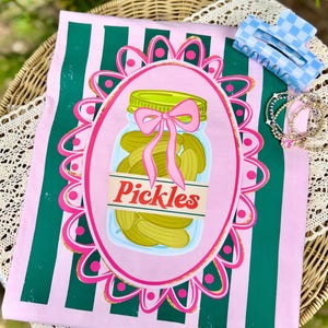 Peut inclure: T-shirt à rayures roses et vertes avec un bocal de cornichons, un nœud rose et le mot "Pickles". Une pince à cheveux à carreaux bleus et blancs et des bracelets sont également visibles.