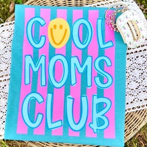 May include: A turquoise t-shirt with pink vertical stripes and the words "COOL MOMS CLUB" in light blue, with a yellow smiley face. A stack of bracelets and a small floral-patterned item are visible.