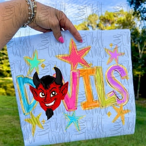 May include: Light blue t-shirt featuring the word "DEVILS" in colorful, outlined letters, with a cartoon devil head and star graphics. The devil has red skin, black horns, and a goatee. The stars are pink, teal, and gold.