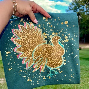 May include: Teal-colored t-shirt featuring a turkey design with gold leopard print feathers, outlined in pink and teal. Gold paint splatters accent the shirt. The shirt is held up against a backdrop of a blue sky and green grass.