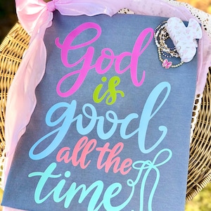 May include: A gray canvas with the words "God is good all the time" in colorful script. A pink bow and sheer ribbon adorn the top left corner. A collection of bracelets and a butterfly charm are on the right.