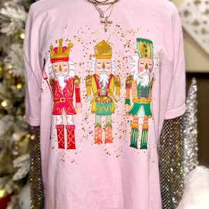 May include: A light pink t-shirt featuring a graphic of three colorful nutcracker soldiers. The nutcrackers are in red, yellow, and green uniforms with gold accents. Gold glitter dots the shirt. The shirt is displayed on a mannequin.