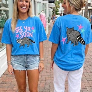 May include: Two people wearing blue t-shirts with a raccoon graphic and the text "SEE YA SOON". The raccoon is grey with a striped tail. The back of the shirt has a raccoon and star graphics. One wears denim shorts, the other white trousers.