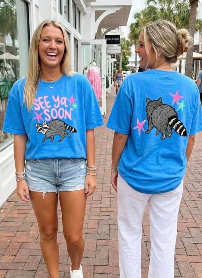 May include: Two people wearing blue t-shirts with a raccoon graphic and the text "SEE YA SOON". The raccoon is grey with a striped tail. The back of the shirt has a raccoon and star graphics. One wears denim shorts, the other white trousers.