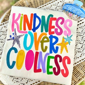 May include: Cream-colored t-shirt with the words "KINDNESS OVER COOLNESS" in vibrant, multicolored block letters. The design includes star and smiley face graphics. A blue hair clip and bracelets are in the upper right corner.