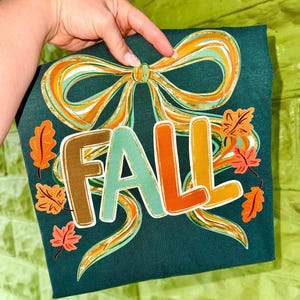 May include: Dark teal t-shirt with a colourful "FALL" graphic. The design features a large bow and autumn leaves in shades of orange, teal, and brown. The word "FALL" is in a playful, layered font.