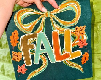 May include: Dark teal t-shirt with a colorful "FALL" graphic. The design features a large bow and autumn leaves in shades of orange, teal, and brown. The word "FALL" is in a playful, layered font.