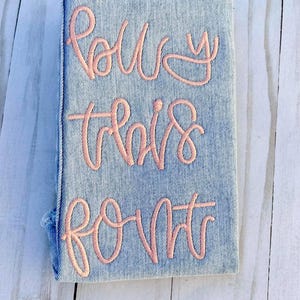 May include: A denim-covered notebook with the words "Follow this font" embroidered in a light pink script. The notebook has a rectangular shape and a slightly distressed appearance, with visible stitching along the spine.