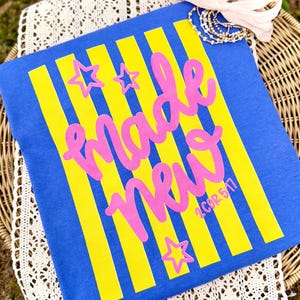 May include: Blue t-shirt with yellow vertical stripes and the words "made new" in pink script. Pink star shapes are scattered around the text. The text "2 COR 5:17" is in the lower right corner.