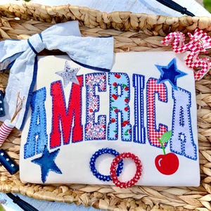 Könnte beinhalten: Ein cremefarbenes T-Shirt mit dem Wort "AMERICA" in roten, blauen und gemusterten Buchstaben. Das Design umfasst eine Kirsche, Sterne und zwei Haargummis. Das Shirt wird in einem Weidenkorb mit einer weißen Schleife präsentiert.